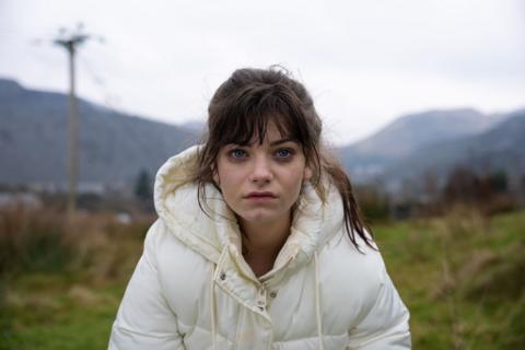 A young woman with brown hair in a ponytail and wearing a white puffer coat stares at the camera with a sad expression
