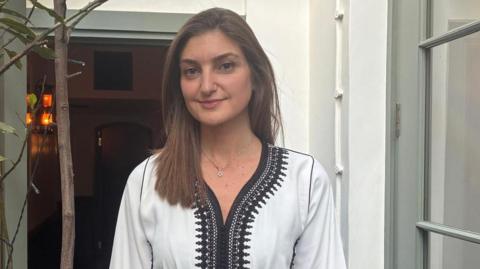 Stephanie Laurent is seen wearing a white kaftan with black embroidery around the V-neckline, smiling for a portrait in a doorway.
