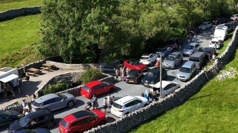Parked cars lining with side of the road at Gordale Scar with pedestrians walking in the road. Some other cars are also driving down the middle of the road.