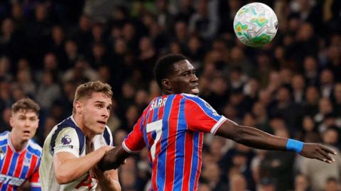 Ismaila Sarr seen from waist up in a Crystal Palace home jersey with red and blue vertical stripes competes for a bouncing ball with Spurs defender Micky van de Ven, who is pulling at Sarr's rear arm. The ball is high up at the top of the image on the right-hand side, with Sarr tilting up his face to look at it as he runs from left to right across the image. A Palace team-mate is visible in the bottom left corner while in the background is a packed crowd
