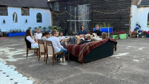 Nine people sat on wooden chairs and a sofa in the middle of a courtyard. 