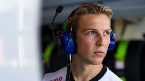 A man with blond hair pushed back wearing a white t-shirt looks to the right of the picture. He's wearing blue over-ear headphones with a microphone attachment angled upwards