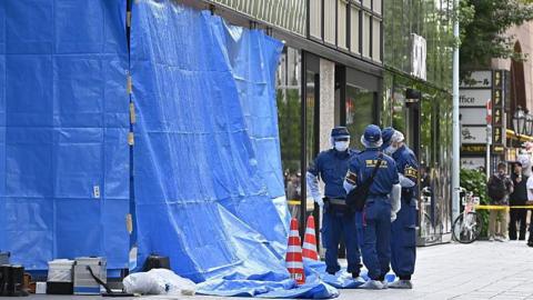 Three people wearing a navy blue uniform and protective gear stand on a street in front of a large shopping centre. Next to them is a large blue tarp sheet with two red and white cones. Behind them is a yellow emergency cordon.