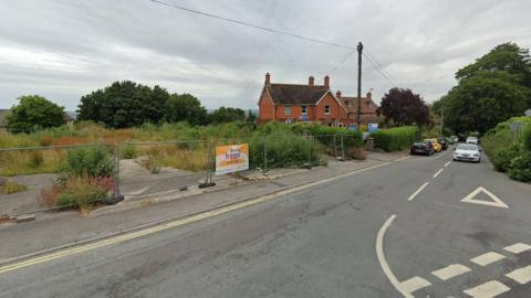 A google street view image of an overgrown site of land, with large metal barriers protecting it. A large red brick house is visible in the background and cars can be seen on road beside it. 