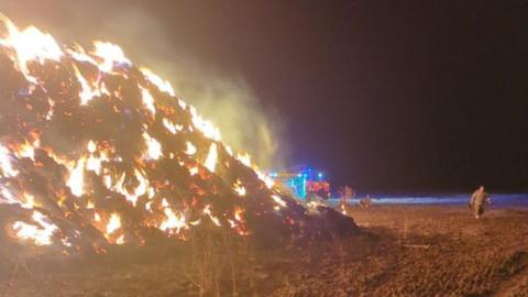 A large haystack fire with two firefighters to the right of the photo and a fire engine in the background