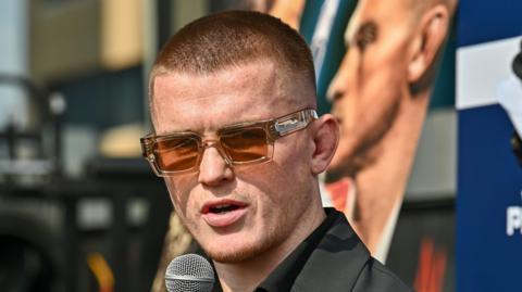 Paul Hughes in sunglasses speaks on a microphone