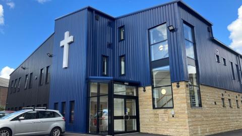 A large building made from stone and corrugated iron with a silver car parked outside