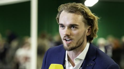 Tom Coyd looks into the distance. He is holding a microphone and speaking