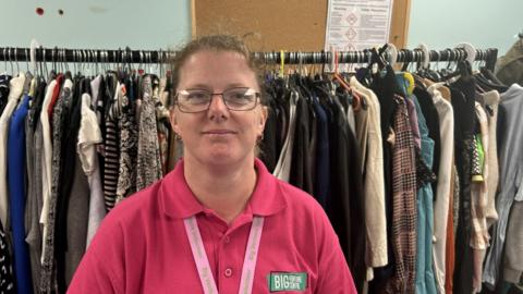 Jane Platt in a bright pink polo shirt. It says Big Venture Centre. Her lanyard says the same. She has glasses and is smiling. There are lots of clothes on a big rack behind her. 