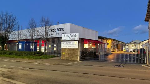 The outside of the Swallowfield site, which is a low square building behind a large fence. The KDC/One logo is visible on the building and on the gate. 