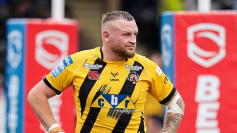 Brad Singleton in a yellow-and-black Castleford shirt with his hands on his hips and the red rugby posts behind him