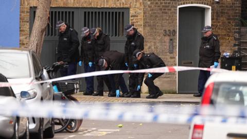 Seven uniformed police officers are investigating the ground and pavement area. They are wearing black Met caps and blue gloves and stood behind police tape. 