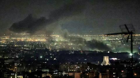 Smoke plumes billow from the site of airstrikes near Azadi Tower in western Tehran on 10 March. The night sky and smoke are black. Below, lights are on in buildings and a tower is lit up.