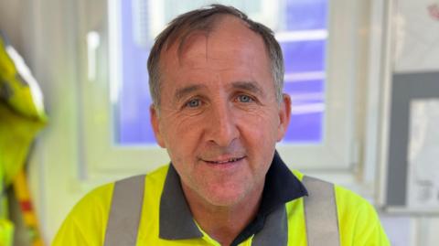 Man in a high-visibility yellow Taylor Wimpey shirt sits in an office chair, smiling at the camera, with a window, hanging work jacket, and site plans visible in the background.