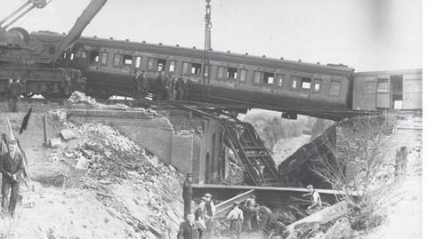 A black and white photo shows a steam train carriage suspended by a crane over a ravine. The mangled wreckage of the railway track on the bridge lies below the carriage. Men stand in the wreckage searching for survivors.