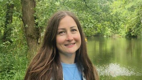 A lady with long, straight brown hair, wearing a light blue top, standing by the side of a large stream and in front of a mature tree, partly hanging over the water.