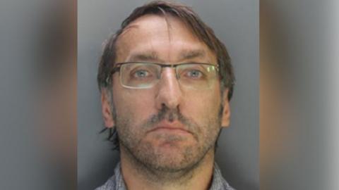 Custody image of Nicholas Stubley who has grey hair and is wearing glasses and a grey and white shirt.