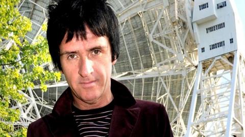 Johnny Marr, with a burgundy velvet jacket and black and white striped top, looks into the camera in front of the Lovell Telescope