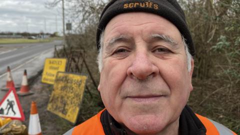 Mark Fishpool, 68, has grey hair around his ears, he is wearing a black beany hat with a high-viz orange jacket. Behind him are a selection of old road signs, stands for them, bollards, sandbags and safety lamps.