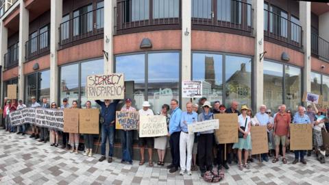 A line of protestors outside a council building, which has high glass windows. They are holding placards and signs saying: "No gas plant dioxins" and the words "Secrecy Transparency", with the word "secrecy" crossed out.