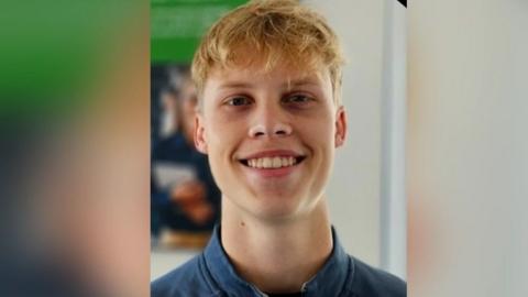 Sebastian Sailes smiles at the camera. He is wearing a blue shirt and has blonde curly hair. 