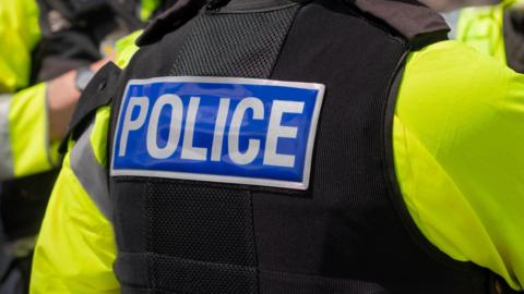 Police officer pictured from behind, wearing a high vis yellow jacket and a black vest with the word 'police' across the back. 