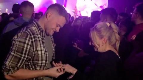 A man with short hair and a chequed brown shirt putting a ring on the finger of a woman with blonde hair tied back behind her head and a black top in a crowded area with a large pink display screen in the background