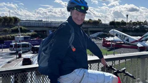 Man wearing a cycle helmet, dark coloured fleece and knee length shorts and a rucksack posed sitting on a bicycle on a pedestrian bridge overlooking a roundabout, with cars and an aeroplane in the background