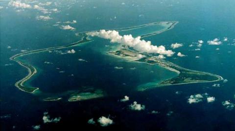 An aerial view of Diego Garcia, the largest island in the Chagos archipelago. Fluffy white clouds are above the island, which is surrounded by deep blue water.