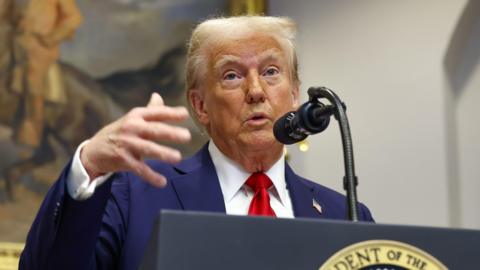 Donald Trump in a blue suit and red tie gestures at a podium