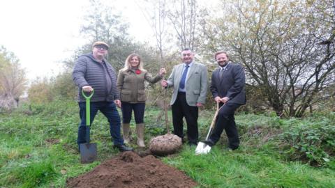Three men and a woman stand on a patch of green grass behind a mound of brown earth. In the centre, a woman with blond hair, dressed in a green jacket, dark trousers and grey wellies, holds a bare tree sapling with a man standing next to her, who wears a grey blazer, black trousers, a pale shirt and a blue tie. To the left, a man wearing a flat cap, blue coat and blue jeans holds a spade with a bright green handle. To the right, a man with short brown hair and a beard, dressed in a dark suit, poses with his foot on a spade as if preparing to dig.