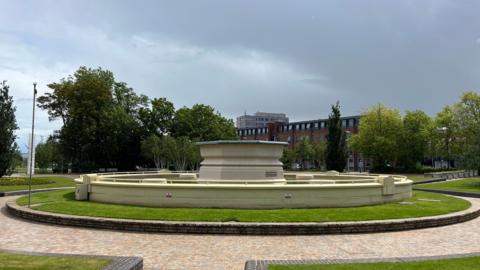 A light yellow brick fountain, without any water coming out. It is surrounded by landscaped gardens and paving. The sun is shining on the fountain but the sky is full of grey clouds.