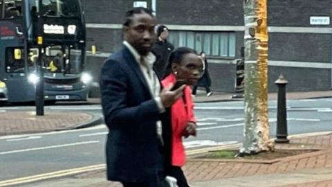 A slightly blurry image of a man and a woman in formal wear, with the man wearing a dark suit and white shirt, and the woman wearing a pink jacket. They are on the street and a bus can be seen on the road behind them. Both are looking at the camera, with the woman leaning forwards to see the camera.