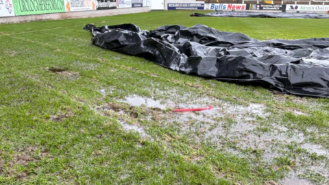 General view of a waterlogged football pitch with some black tarpaulin on it. At the top of the picture a series of advertising hoardings can be seen on the edge of the pitch.