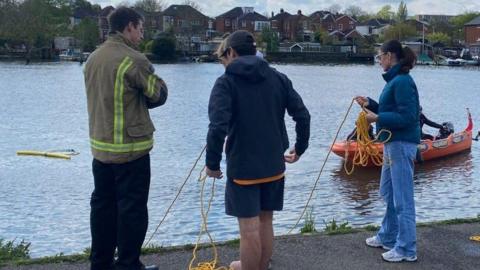 A emergency services worker, in a high-vis jacket, stood next to three people who are testing the bright yellow ropes that have been put in the river. There is also a RIB and a yellow float visible on the water in the background.