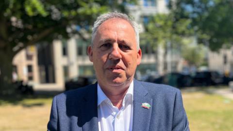 Constable Andy Jehan is a man with grey hair. He is looking into the camera. He is standing in Parade Park, with the government building out-of-focus behind him. He is wearing a grey suit and white shirt.