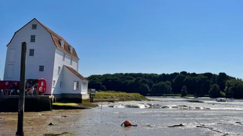 On the left is a large white mill building.  It's on the edge of the river and is around four or five storeys high. It's white and is made up of horizontal wooden slats.  There are four small windows on the side of the building and a further five on the front with two windows in the tiled roof.  The tide is out and the mud of the river bed is visible. There are a lot of trees in the background and a blue, cloudless sky.