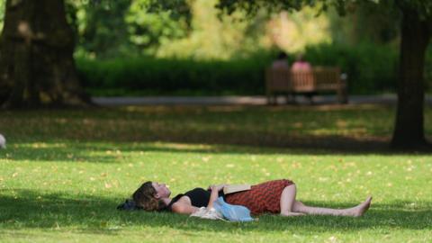 A woman lies down in a park on the grass with a book on her lap 