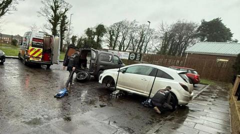 A number of cars in various colours are parked in a car park. A work van is seen in the background as workers attempt to fix tyres.