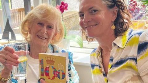 An elderly woman with blonde hair holding a glass and a 100th birthday card. Next to her is younger woman with brown hair wearing a white, blue and yellow shirt.