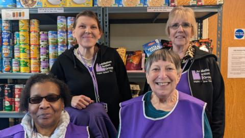 Four women photographed in front of a shelving unit stacked with food items, mostly tins. They are wearing purple tabards with "Jubilation Action Group" logos on them.