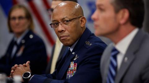 CQ Brown in navy blue uniform and glasses, with hands folded looks over at Pete Hegseth in grey suit