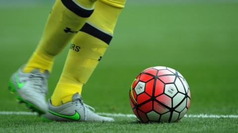 A person's feet wearing football socks and boots stand next to a football on a green pitch.