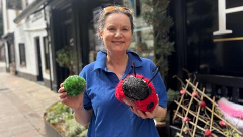 A woman, wearing a blue t-shirt and sunglasses on her head. She is holding a knitted Brussels sprout and knitted ladybird. 