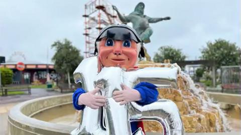 The Jolly Fisherman mascot holding silver balloons in front of the Jolly Fisherman statue and fountain