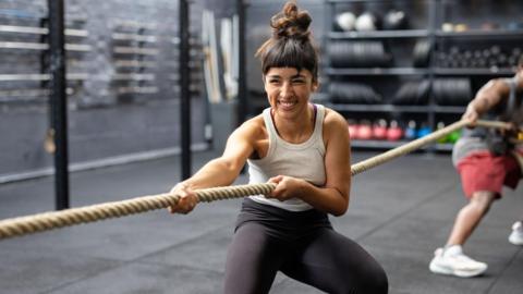 A young woman wearing black leggings and a sleeveless top, with dark hair tied up, pulls hard on a tug-of-war rope in a gym with a grey floor. Shelves of weights and are seen in the background, as well as a man (blurred) holding the same rope, wearing red shorts.