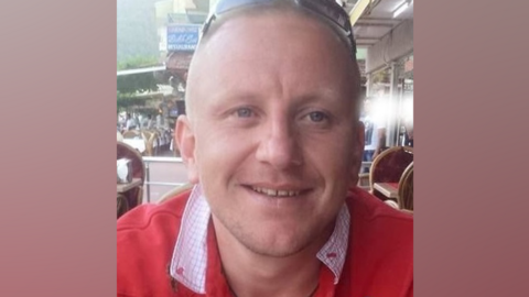 A man with cropped fair hair and wearing a red jumper and sunglasses on his head smiles at the camera. He is sitting on a restaurant terrace. 