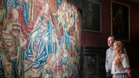 A man and woman are looking at a Tudor tapestry which depicts a scene from the Old Testament Book of Esther and is blue and red in colour.