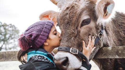 A woman in a woolly hat embraces a cow which has its head over a fence