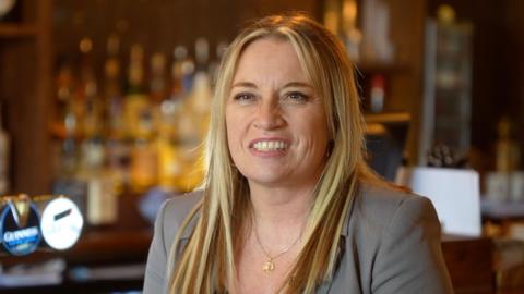 A head and shoulders image of Rachael Howlett, manager of the Claydon Greyhound pub, smiling while looking past the camera. She is wearing a grey blazer and a gold necklace. She is sitting in front of a pub bar.